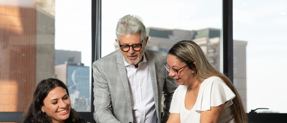 Consultores de contabilidade digital analisando dados financeiros com cliente. Homem de paletó cinza e duas mulheres sorridentes olham para uma tela (não visível), demonstrando suporte humanizado e gestão empresarial eficiente em escritório moderno.