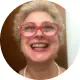 Foto circular de uma mulher loira sorridente, com cabelos curtos e óculos de armação vermelha vibrante. Ela usa um colar de pérolas e demonstra uma expressão de grande alegria e satisfação. O fundo é neutro.