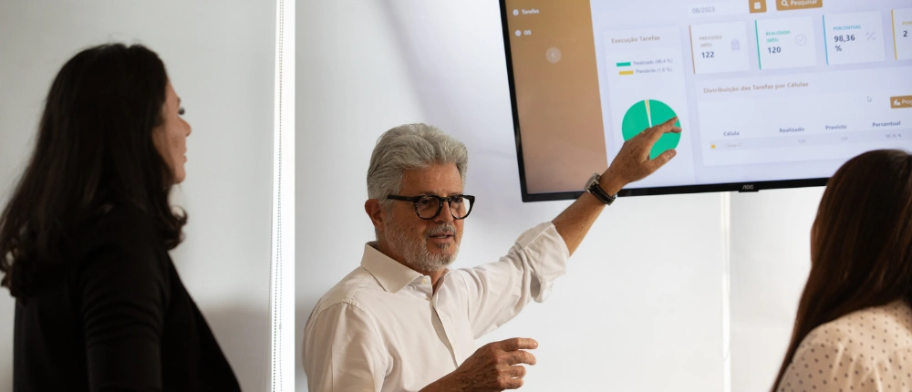 Reunião de análise de resultados financeiros com especialistas em contabilidade digital. Homem de camisa branca e óculos mostra dashboard de controle de tarefas e métricas empresariais em monitor para equipe, demonstrando contabilidade consultiva e eficiência financeira.
