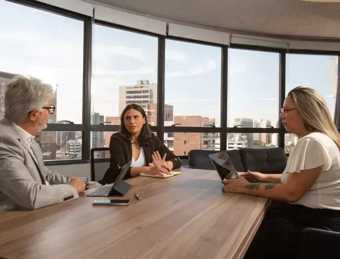Três profissionais reunidos em uma sala de escritório com vista para a cidade, conversando ao redor de uma mesa com notebook e bloco de anotações, em um contexto de reunião ou alinhamento estratégico.