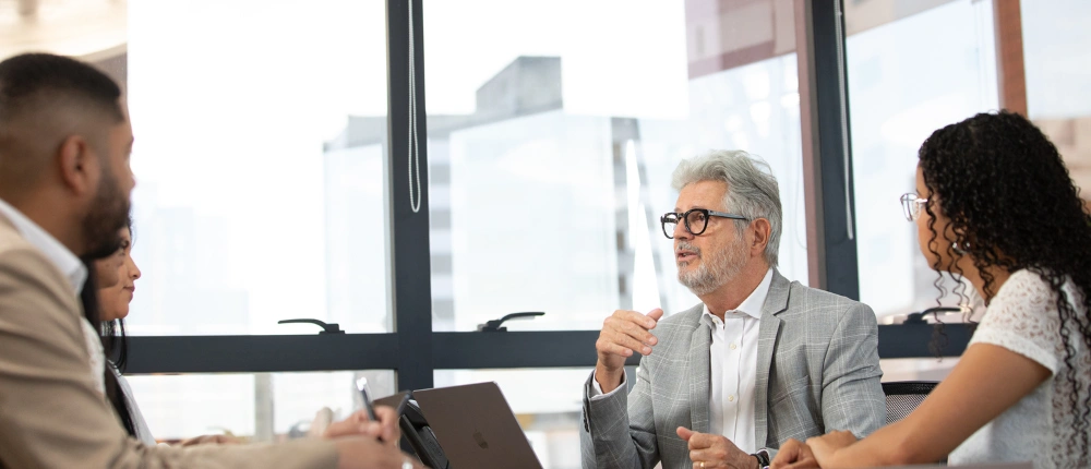 Equipe de contabilidade consultiva em reunião estratégica com clientes. Homem de paletó cinza explica processos de gestão financeira e contabilidade digital em uma mesa de trabalho com computadores.