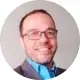 Foto circular de rosto de um homem sorridente com cabelos curtos e óculos de grau de armação escura. Ele veste uma blusa de gola alta azul por baixo de um paletó cinza. O fundo é neutro. Imagem transmite profissionalismo e confiança.