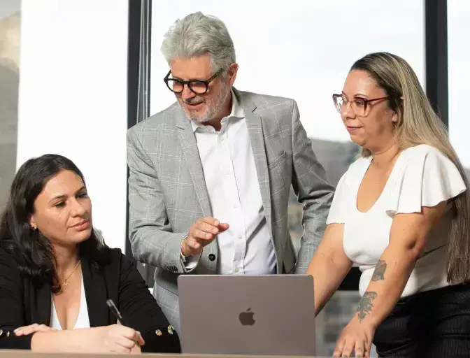 Três profissionais colaborando em um escritório moderno. Um homem de cabelos grisalhos e terno cinza está de pé, usando óculos e apontando para a tela de um laptop aberto. Ao seu lado, uma mulher com óculos e blusa branca observa atentamente, enquanto outra mulher, sentada à esquerda com um blazer preto, parece fazer anotações. O ambiente é bem iluminado, sugerindo uma reunião de trabalho ou consultoria.